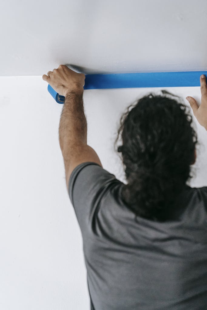Adult applying blue tape to ceiling for painting, focused on precision.