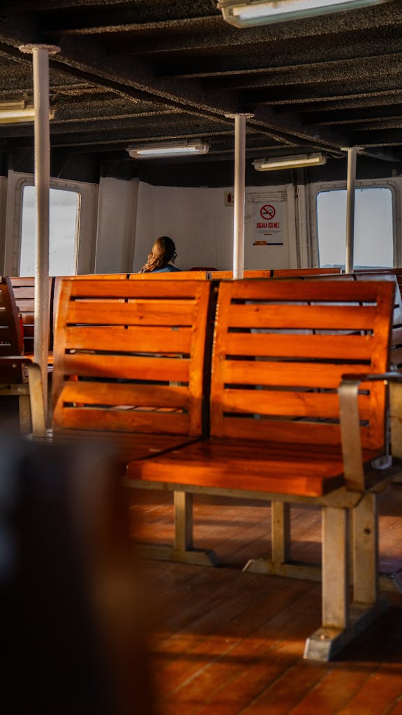 Sunlit empty ferry interior featuring wooden benches and a distant passenger.